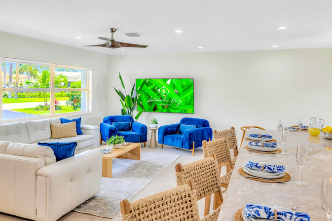 Sunlit coastal living room with white leather sofa, two blue velvet armchairs, woven rattan barstools at a marble island, tropical plants and large window with palm trees outside.