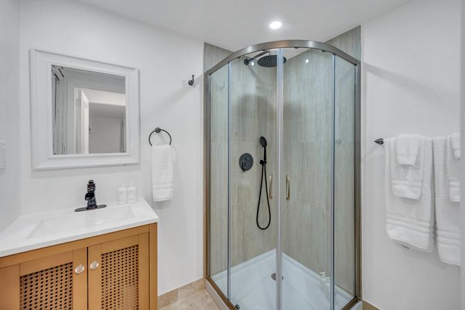 Modern white bathroom with a curved corner glass shower featuring a rain showerhead and handheld sprayer, light beige tiled walls, white sink and framed mirror, wooden vanity cabinet with lattice doors and neatly folded white towels.