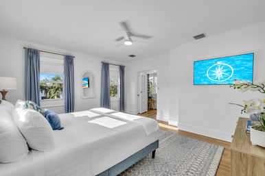 Sunlit coastal-style bedroom with white bedding, blue curtains, ceiling fan, hardwood floors and wall-mounted TV showing a compass over water; palm trees visible through the windows.