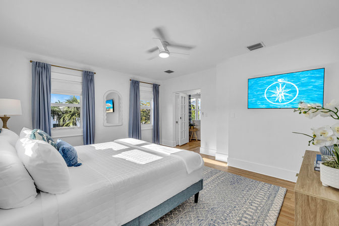 Sunlit coastal-style bedroom with white bedding, blue curtains, ceiling fan, hardwood floors and wall-mounted TV showing a compass over water; palm trees visible through the windows.