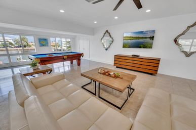 Bright waterfront condo game room with cream leather sectional, wooden coffee table with charcuterie, billiards table by large canal-view windows, wall-mounted TV and decorative mirrors.