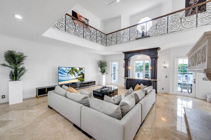 Spacious luxury waterfront living room with double-height ceiling and ornate wrought-iron balcony, polished marble floors, oversized light-gray sectional around a black coffee table, flat-screen TV showing a tropical beach, dark wood wet bar, potted palms and French doors opening to a dock.