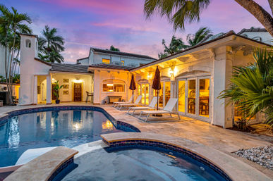 Luxury tropical villa patio at sunset — Mediterranean-style home with warm interior lights, palm trees, pool and spa, plus sun loungers and umbrellas