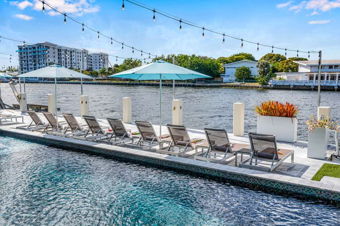 Riverside waterfront pool deck on a sunny day with a row of lounge chairs, turquoise umbrellas, string lights overhead, and modern buildings across the water.