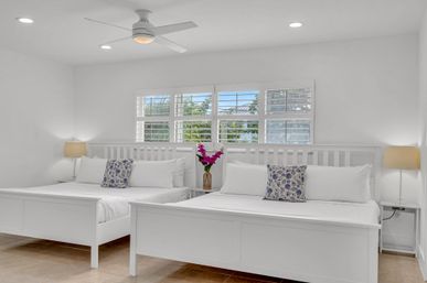 Sunlit coastal guestroom with two white beds, ceiling fan, plantation shutters revealing palm-tree view, blue floral pillows and pink flowers on a bedside table.