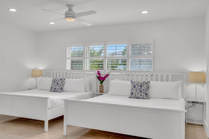 Sunlit coastal guestroom with two white beds, ceiling fan, plantation shutters revealing palm-tree view, blue floral pillows and pink flowers on a bedside table.