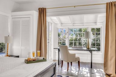 Sunlit bedroom with a windowed home office — desk and beige chair by mullioned windows overlooking a leafy garden, bed with breakfast tray and neutral curtains.