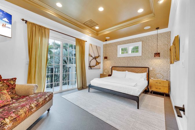 Bright modern tropical bedroom with king-size bed, textured accent wall, gold tray ceiling, pendant lights, sliding glass doors to a balcony with palm trees and a patterned chaise lounge.