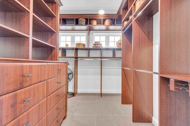 Bright walk-in closet with warm wood built-in drawers, open shelving, hanging rods and cubby storage under small windows, tiled floor and a standing fan tucked in a corner.