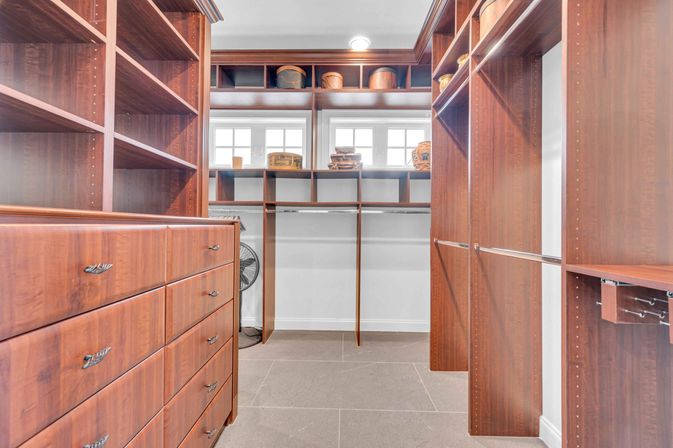 Bright walk-in closet with warm wood built-in drawers, open shelving, hanging rods and cubby storage under small windows, tiled floor and a standing fan tucked in a corner.