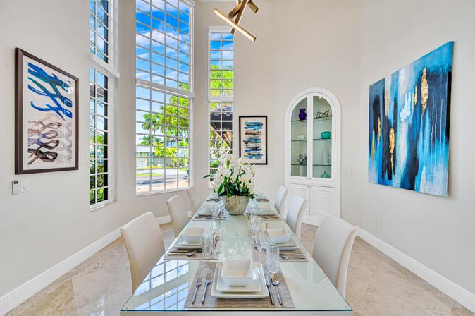 Bright modern coastal dining room with glass-top table set for eight, white upholstered chairs, floor-to-ceiling windows showing palm trees and waterfront, blue abstract paintings, built-in glass cabinet, orchid centerpiece.