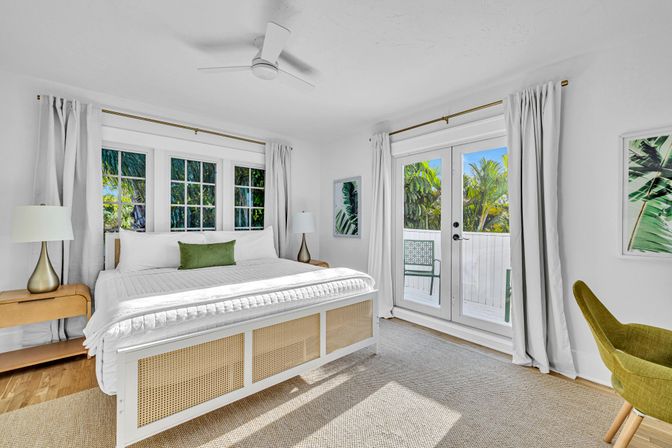 Sunlit tropical bedroom with a large white bed and woven footboard, green accent pillow, bedside lamps, ceiling fan, and French doors opening to a balcony framed by lush palm foliage.