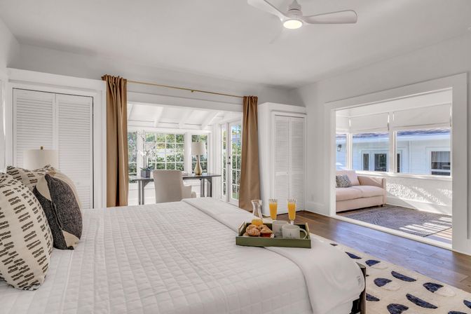 Bright sunlit bedroom with white quilted bed, patterned pillows, breakfast tray with croissants and orange juice, desk in a glass-enclosed sunroom and adjoining cozy seating area on hardwood floors