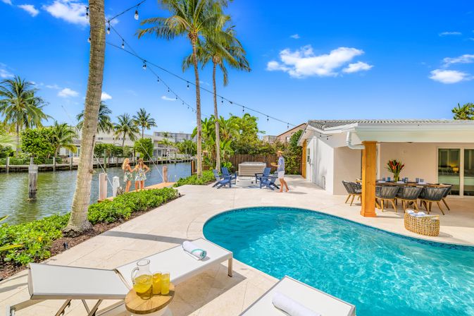 Sunny canal-front tropical backyard with curved blue pool, palm trees and string lights, lounge chairs with lemonade, outdoor dining area and dock along a calm waterfront — vacation-style patio.