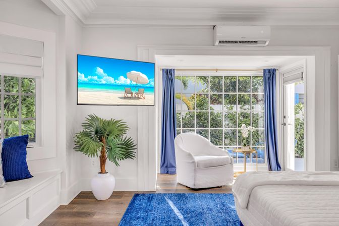 Sunlit coastal bedroom with white bed and armchair, large grid windows and glass door to a leafy patio, blue rug and curtains, potted palm, and wall-mounted TV showing a beach scene — bright beachy vibe.