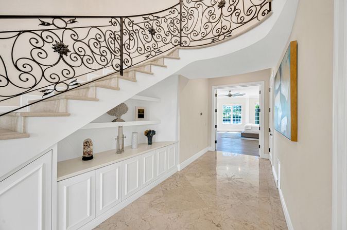 Elegant marble-floored foyer with curved wrought-iron balustrade staircase, built-in white display shelves and cabinetry under the stairs, leading to a bright sunlit living room with large windows.