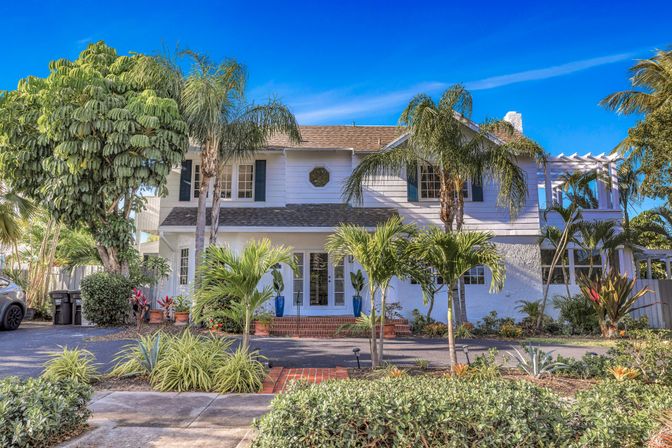 Sunny two-story white coastal house framed by palm trees and tropical landscaping, brick steps and potted plants leading to the front doors under a bright blue sky