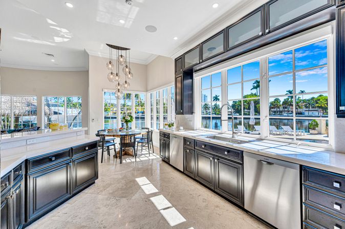 Sunny waterfront kitchen with dark wood cabinets and marble countertops, large windows overlooking a canal with palm trees and a dock, stainless-steel appliances, breakfast nook and modern pendant lights.