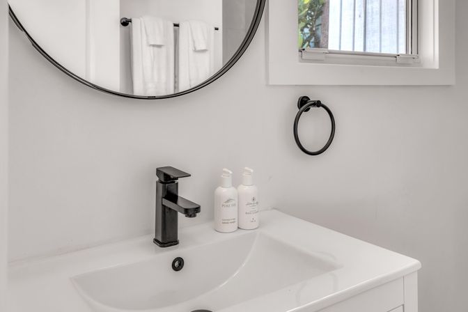 Minimalist white bathroom vanity with matte-black faucet and towel ring, round mirror reflecting folded white towels and a small window with natural light.