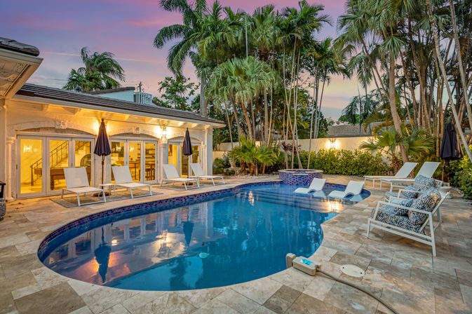 Twilight tropical backyard with a curved blue-tiled pool and raised spa, stone tile patio lined with lounge chairs and umbrellas, palm trees and glass doors opening to a villa.