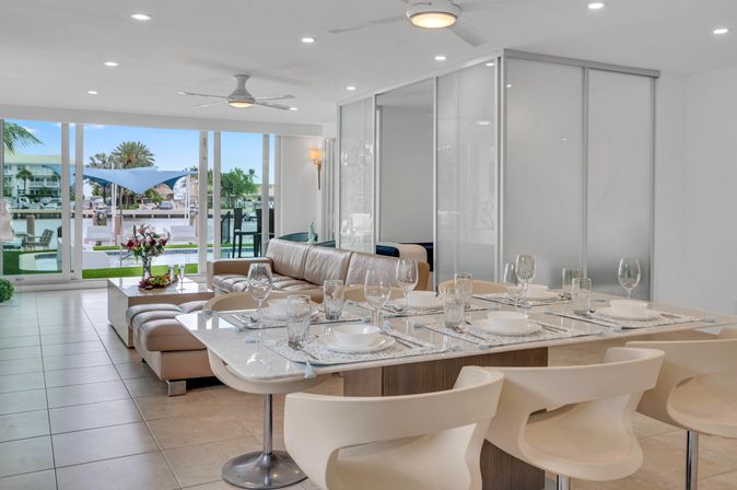 Bright modern waterfront condo open-plan living and dining area with table set for six, beige leather sofa and sliding glass doors opening to a marina and palm-tree dock view.