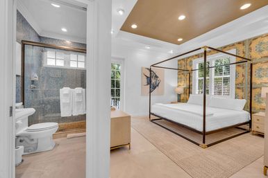 Bright tropical master bedroom with metal four-poster canopy bed, patterned gold wallpaper, plantation-shuttered windows with palm views, and an adjacent en-suite bathroom featuring a mosaic-tiled shower, white towels, and toilet.