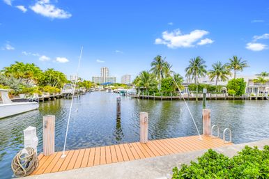 Sunny tropical waterfront canal with a wooden dock and boat slips, palm trees and waterfront homes, and a distant coastal city skyline under a bright blue sky.
