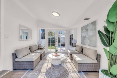 Bright, airy sunroom with wicker sofas and beige cushions facing glass French doors to a sunny backyard, round white coffee table, patterned rug, tropical potted plant and modern wall art.