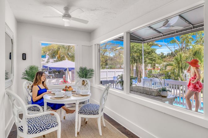 Sunlit coastal breakfast nook with white furniture, woman in a blue dress dining, screened deck, palm trees and pool with a child in a red sunhat