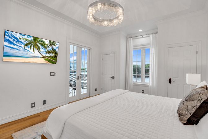 Bright luxury waterfront bedroom with white quilted bed, crystal chandelier, wall-mounted TV showing a tropical beach, French doors to balcony overlooking marina and water.