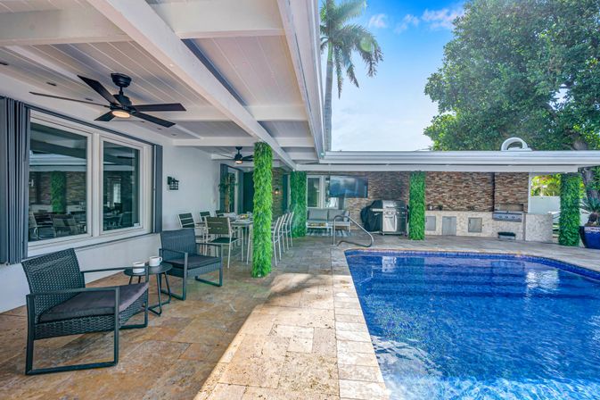 Covered backyard patio with wicker seating, dining area, ceiling fans and outdoor kitchen with grill beside a blue-tiled swimming pool and palm trees