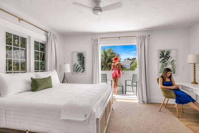 Bright tropical vacation bedroom with white linens and green accent pillow, sliding doors opening to a balcony framed by palm trees and blue sky; a person in a red sunhat stands on the balcony while another sits at a desk in a blue dress, airy modern decor.