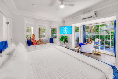 Bright tropical vacation rental bedroom — sunlit coastal suite with large white bed, window seat with blue pillows, wall-mounted TV, ceiling fan, and two guests relaxing by floor-to-ceiling windows opening to a leafy patio
