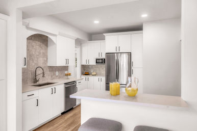 Bright modern white open-concept kitchen with stainless steel refrigerator and dishwasher, quartz countertops, stone subway-tile backsplash, breakfast bar with pitcher and glass of orange juice, gray stools and light wood floors.