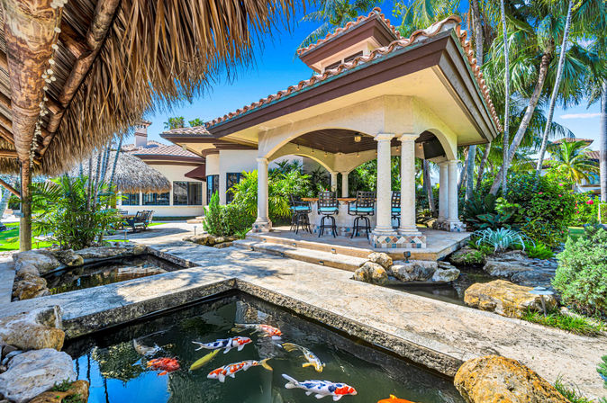 Luxury tropical backyard with colorful koi pond, stone walkways, thatched palapa, Mediterranean-style covered pavilion with columns and outdoor bar seating, tiled roof, palm trees and lush landscaping.