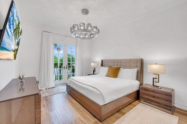 Bright coastal bedroom with king bed and tufted headboard, wooden nightstands and dresser, modern glass chandelier and wall TV, plus French doors opening to a balcony with palm trees and turquoise water beyond.