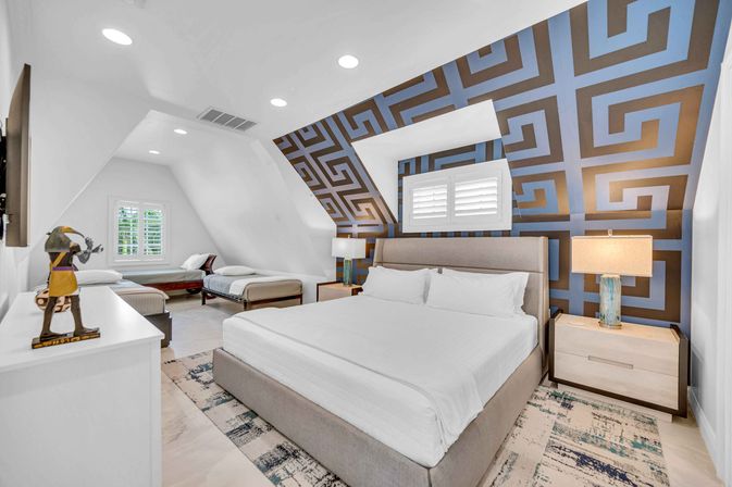 Bright attic-style bedroom with a king bed, two twin beds in a tucked-in alcove, bold blue geometric accent wall, white plantation shutters, modern nightstands with lamps, patterned rug and recessed ceiling lights.