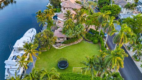 Aerial view of a sunlit waterfront tropical estate on a calm canal — luxury villa surrounded by palm trees, private dock with yacht, and a large lawn with trampoline and soccer goal