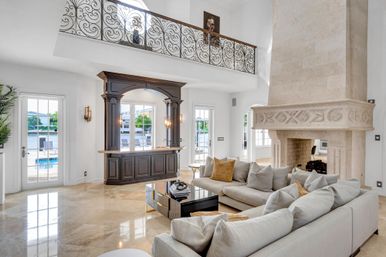 Sunlit luxury waterfront living room with polished marble floors, oversized carved stone fireplace, L-shaped beige sofa, glass coffee table, ornate wrought-iron balcony and dark wood wet bar overlooking a pool and water view.