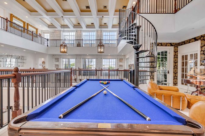 Bright two-story luxury home game room with electric-blue pool table and crossed cues, spiral iron staircase, leather seating, and sunlit mezzanine balcony