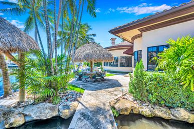 Sunny tropical waterfront patio with thatched palapa dining area, swivel chairs, tall palm trees, lush landscaping and a canal beside a Mediterranean-style luxury home