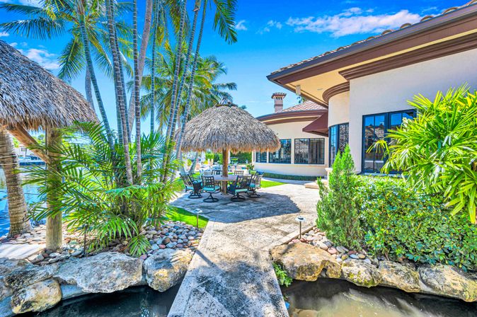 Sunny tropical waterfront patio with thatched palapa dining area, swivel chairs, tall palm trees, lush landscaping and a canal beside a Mediterranean-style luxury home