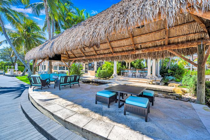 Tropical waterfront patio with a large thatched palapa, turquoise‑cushioned outdoor seating, shaded dining area, palm trees and a stone walkway under a bright blue sky.