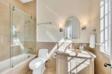 Sun-drenched elegant bathroom with beige marble tiles, glass sliding bathtub enclosure, white toilet, carved vanity topped with an arched mirror, wall sconces and a towel ring by the window.