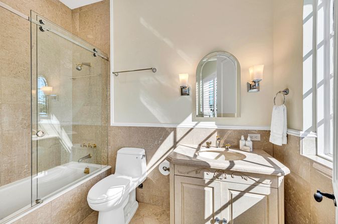 Sun-drenched elegant bathroom with beige marble tiles, glass sliding bathtub enclosure, white toilet, carved vanity topped with an arched mirror, wall sconces and a towel ring by the window.