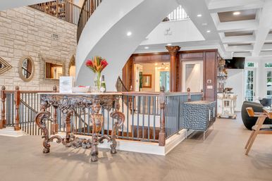 Bright luxury foyer with ornate carved wooden console table topped with flowers and champagne, sweeping arch, iron-railed staircase, stone accent wall and coffered ceiling.