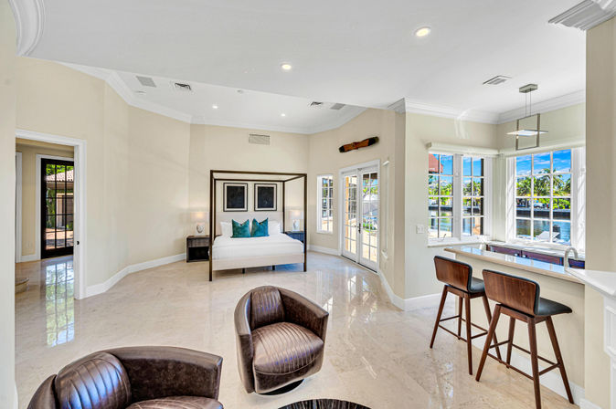 Sunny coastal waterfront master suite with four-poster bed, marble floors, leather swivel chairs, breakfast bar with two stools, and French doors and large windows overlooking a canal and palm trees.