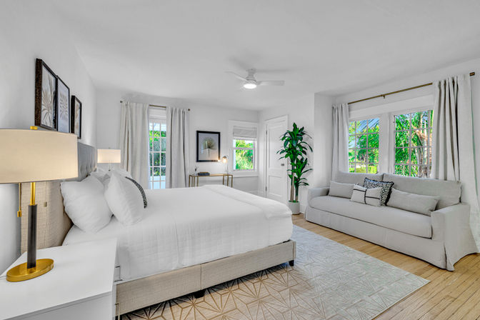 Airy sunlit coastal-style bedroom with crisp white bedding, upholstered headboard, light-gray sofa, large windows framing lush tropical greenery, a potted plant, bedside lamps and light wood floors.