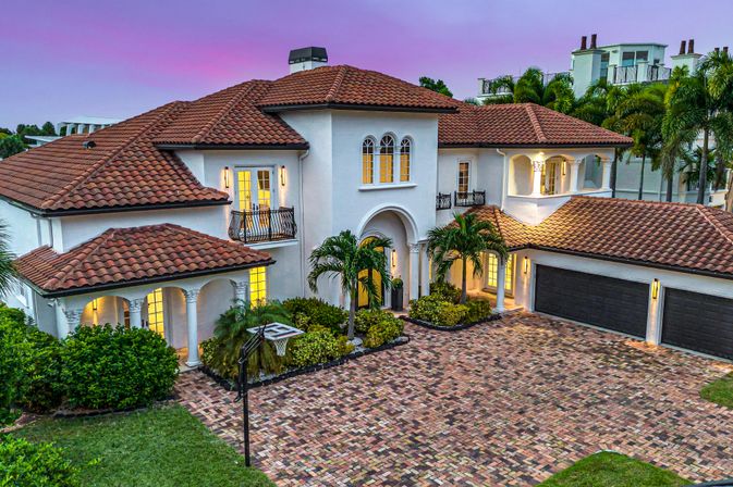 Mediterranean-style luxury home at twilight with red clay tile roof, white stucco, arched windows, wrought-iron balconies, palm trees, brick driveway, three-car garage and a basketball hoop