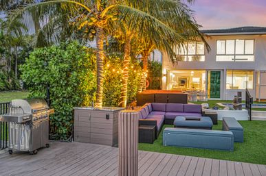 Cozy tropical backyard patio at dusk with illuminated palm trees wrapped in string lights, stainless steel gas grill on a wooden deck, modular purple outdoor sectional on artificial turf, and a modern two-story home in the background.